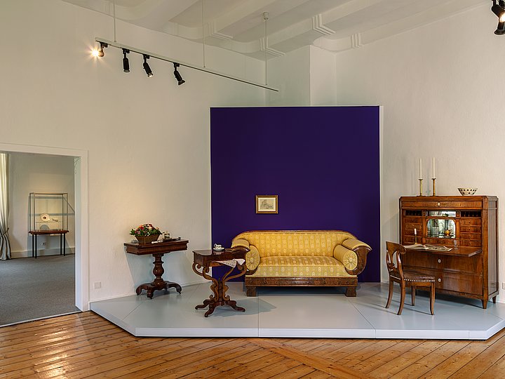Wohnzimmer-Arrangement mit Sofa und Sekretär vor lila Wandfeld